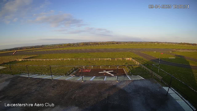 A wide view of an open airfield under a partly sunny sky. The foreground shows the edge of a building with a flat roof, and there is a helipad marked with a white cross on a brown surface. In the background, there are green fields and a long runway leading into the distance, bordered by a wooden fence. A windsock is visible near the runway, indicating wind direction. The time and date are displayed in the top right corner.