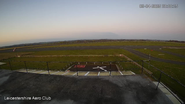 A view from a webcam showcasing an airfield at twilight. The foreground features a flat rooftop with a railing and an aerial view of a small runway made of asphalt, with grassy areas on either side. There is a large white X symbol on the ground, indicating a helipad area. In the distance, rolling hills are visible under a pastel sky. The image includes a timestamp in the upper right corner, indicating the date and time.