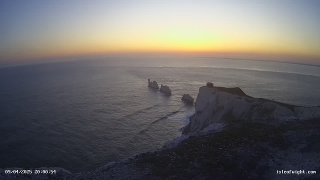 A scenic view of the sea and cliffs during sunset. The sky transitions from soft blue to warm orange and yellow hues as the sun sets on the horizon. In the foreground, there are white chalk cliffs with a small structure on top. Three rock formations are partially submerged in the water, creating gentle waves that ripple towards the shore. The overall atmosphere is calm and serene.