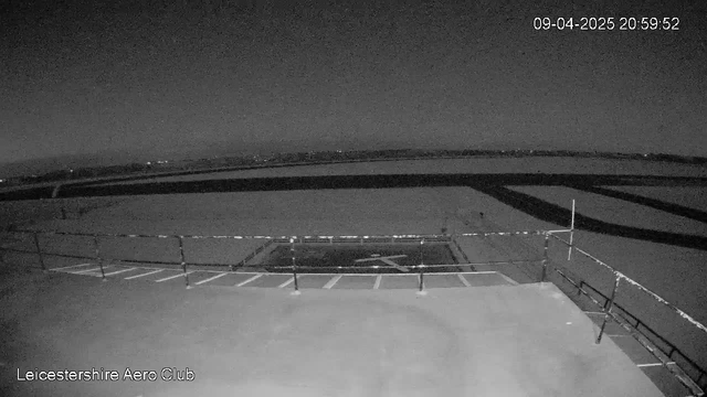 A dark, nighttime view of an airstrip from the perspective of a webcam. In the foreground, there is a flat surface with a marked "X" indicating a landing spot, bordered by white lines. A railing is visible along the edge, and the background features a wide expanse of land with subtle outlines of trees or buildings in the distance, suggesting a rural setting. The scene is largely shadowy with limited visibility, illuminated only by faint lights in the far background. The timestamp is displayed in the upper right corner, showing the date and time.