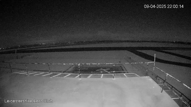 A dimly lit view from a webcam positioned on a rooftop, showing a large, flat surface likely intended for landing or parking aircraft. In the foreground, there is a white marking in the shape of an "H" for a helicopter landing pad. Beyond the landing area, a dark, expansive field stretches out, with faint outlines of grass and possibly trees. The horizon is visible in the distance, with a few lights twinkling, indicating nearby structures or roads. The scene conveys a quiet and tranquil nighttime atmosphere. The timestamp in the corner reads "09-04-2025 22:00:14" and the text "Leicestershire Aero Club" is displayed at the bottom.
