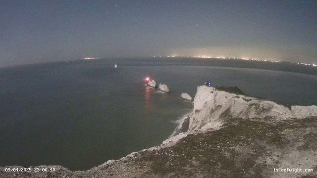 A shoreline at night with rocky cliffs and calm waters. There are several jagged rock formations emerging from the water, with a small red light visible on one of the rocks. The cliffs are mostly white with some grassy patches, and in the distance, faint lights from a city can be seen along the horizon. The sky is mostly clear with a few stars visible.