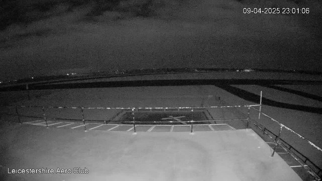 A dark image showing a rooftop view of an airfield. In the foreground, a railing outlines the edge of the roof, and a section of the ground is marked with white lines indicating possible parking spaces. The background is mostly empty with some distant city lights hinting at a horizon under a cloudy night sky. The date and time are displayed in the top right corner, indicating a nighttime setting.