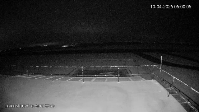 A dark scene captured from a webcam at Leicestershire Aero Club. The foreground shows a flat surface with a railing visible on the right side. There are faint lights in the distance, suggesting an area beyond the site, but the overall scene is mostly dark with minimal details. The time stamp at the top indicates it is 5:00 AM on April 10, 2025.