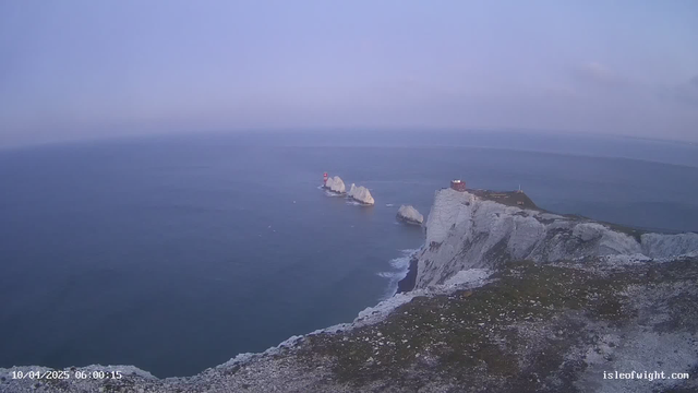A panoramic view of a coastal landscape at dawn. The foreground features white chalk cliffs with patches of grass. In the middle of the image, several rocky outcrops rise from the sea, with a red lighthouse on one of them. The sea extends to the horizon, reflecting a soft blue color, while a light blue sky dominates the background, gradually transitioning from light to darker shades.