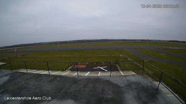 A view from a webcam at the Leicestershire Aero Club, showing a large open space with grassy fields and a tarmac runway. The sky is overcast and grey, with no visible sun. In the foreground, there is a railing, and a faint outline of a white cross marked on the ground. The runway is visible leading into the distance, with areas of grass along the sides. The image includes a timestamp in the top right corner indicating the date and time.