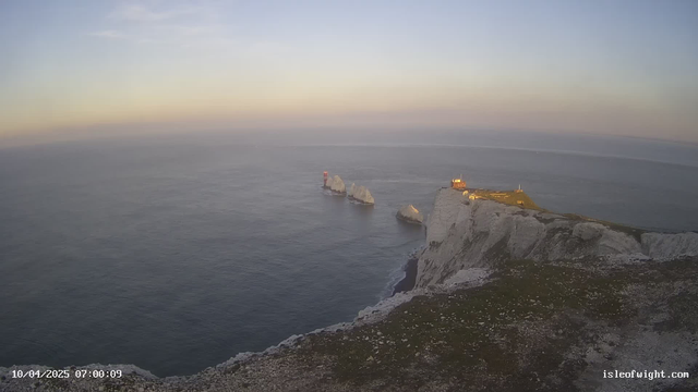 A scenic coastal view features a rocky shoreline with white cliffs on the right side. In the foreground, there is grass and scattered stones. The ocean stretches out to the horizon, which is painted in soft hues of orange and blue from the early morning light. Three jagged rock formations emerge from the water, with a red navigational buoy visible to the left. At the top of the cliffs, a small structure with illuminated windows indicates a lighthouse or lookout point. The sky is mostly clear with a few light clouds, enhancing the tranquil atmosphere.