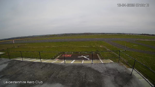 A view from a webcam at Leicestershire Aero Club, capturing a grassy landscape with a runway and scattered markings. In the foreground, there is a flat rooftop surface with a railing. The horizon is mostly cloudy, and the scene appears tranquil, indicating early morning. A red sign is visible on the ground near the center, and a white symbol resembling a large cross is painted on the ground. The runway extends into the distance, bordered by a sparse fence.