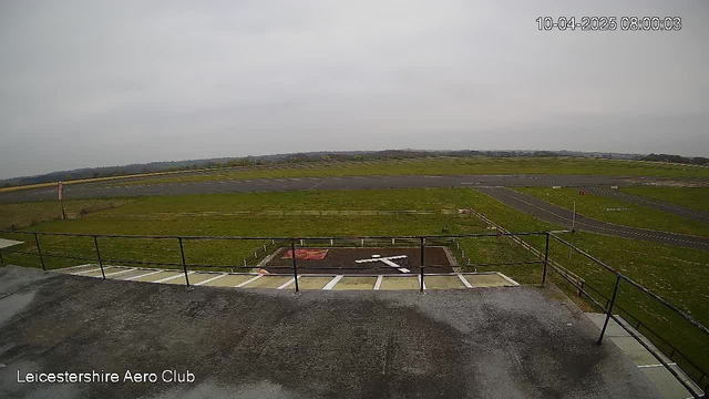 A view from a webcam positioned on a building at the Leicestershire Aero Club. The image shows a large expanse of grassy land with an airstrip running diagonally across the scene. In the foreground, there is a patio area with railing, and a white cross marked on the ground. The sky is cloudy and overcast, indicating a dull day. Various signs and markings can be seen along the edges of the airstrip.