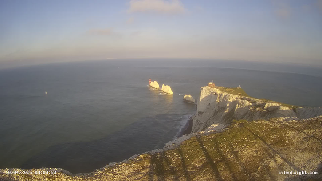 A panoramic view of the sea with gentle waves reflecting the morning light. In the distance, there is a small white sailing boat. Prominent cliffs with white chalk formations rise on the right side of the image, topped with green grass and a few patches of brown earth. At the edge of the cliffs, a red and white lighthouse sits on a rocky outcrop. The sky is mostly clear with a gentle gradient from blue to light gray. The scene conveys a serene coastal atmosphere.