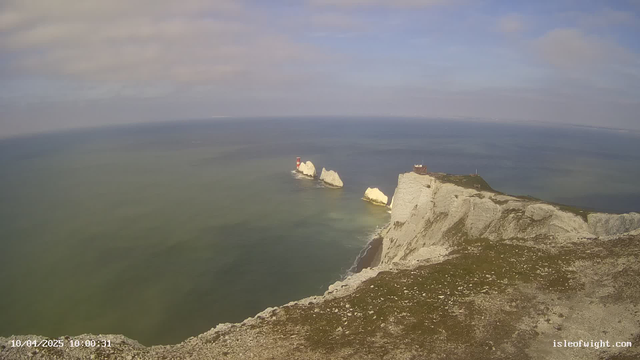 A panoramic view of coastal cliffs meets a calm sea under a mix of blue and cloudy skies. In the distance, two white chalk cliffs rise from the water, with a red and white lighthouse on the left cliff. The foreground features a grassy area with scattered stones, and the shoreline trails off into the water. The scene appears tranquil and natural, with subtle variations in the sea's color.