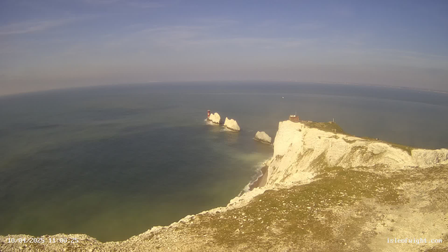 A coastal landscape featuring white chalk cliffs overlooking the ocean. In the distance, there are two distinctive rock formations with a small red lighthouse on one of them. The water is a mix of blue and green hues, with gentle waves lapping against the shore. The sky is mostly clear with some light clouds, and the scene conveys a calm, serene atmosphere.