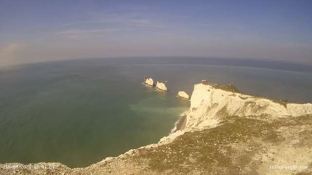 A coastal scene featuring white cliffs rising above a calm turquoise sea. In the background, several rock formations emerge from the water, with a lighthouse visible on one of the rocks. The sky is blue with wispy clouds, and the sun is shining. The foreground shows a grassy area with a rocky texture, leading to the edge of the cliffs.