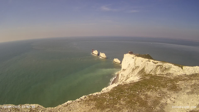 A coastal view featuring chalk cliffs descending into the sea. In the background, there are two prominent white rock formations jutting out of the water, with a small building and a red structure visible on one of the rocks. The sky above is mostly clear with a slight gradient from blue to lighter hues at the horizon. The water appears calm, reflecting various shades of blue and green. In the foreground, the terrain is rocky and grassy, leading up to the cliffs.