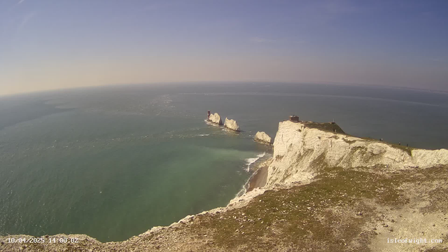 A coastal scene showcasing white chalk cliffs along a shoreline. The ocean is visible in the background with waves gently rolling towards the shore. A few rocky outcrops are present in the water, with one featuring a red structure on top. The sky is clear with some light clouds, and the overall atmosphere appears bright and sunny. There is green grass and rocky terrain at the cliff's edge, and a few people can be seen walking near the top of the cliffs.