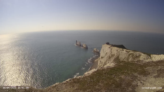 A coastal view with white chalk cliffs extending to the left, overlooking a calm, blue ocean. In the distance, several rocky formations emerge from the water. The sky is mostly clear with a soft hazy horizon. The scene captures a tranquil atmosphere of nature by the sea.