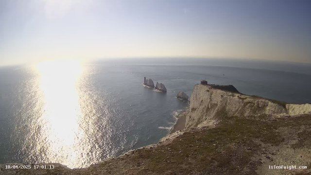 A coastal view during the day, featuring a bright sun reflecting off the water, creating a shimmering effect on the ocean's surface. In the foreground, a grassy cliff edge leads down to a sandy beach. Several large, jagged rock formations jut out from the sea, with one having a structure resembling a lighthouse on top. The sky is clear with a slight haze near the horizon.