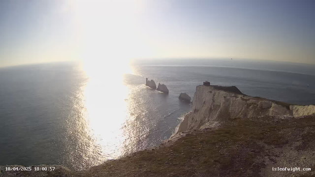 A wide view of the ocean under a bright sun, casting a shimmering reflection on the water's surface. In the foreground, there are grassy cliffs that lead down to the shoreline. To the right, three large rock formations emerge from the water, with a small tower-like structure on top of one. The background features a clear sky transitioning from light blue to a soft yellow hue near the sun. A few boats are visible in the distance on the water.