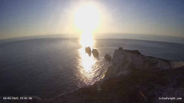 A scenic view of the ocean at sunset. The horizon is bright with the sun lowering, creating a path of shimmering light across the water. In the foreground, jagged rock formations emerge from the sea, while a cliff with grassy areas is visible on the right side. The sky transitions from blue to light hues, indicating the evening. The date and time are displayed at the bottom left corner.