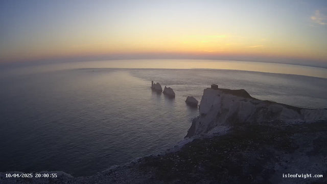 A serene seascape at sunset, featuring calm waters reflecting soft hues of orange and blue. In the foreground, white chalk cliffs rise sharply from the water, with three distinctive rock formations jutting out from the sea. A small structure stands at the top of the cliffs, while the horizon is lined with a faint outline of land in the distance. The sky transitions from light orange near the horizon to a deeper blue above, creating a tranquil and atmospheric scene.