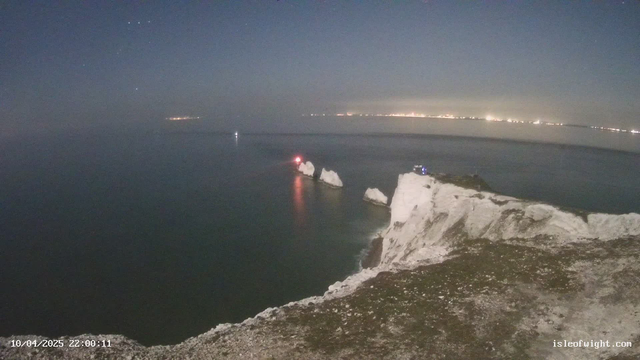 A nighttime view of a rocky coastline with white cliffs. In the foreground, the cliff face is prominent, showing jagged rock formations. The ocean is calm and appears dark with reflections from the water. In the distance, a few white rock outcrops are visible with a red light on one of them, likely a lighthouse or a buoy. There are faint lights from a distant shoreline on the horizon, suggesting a town or city. The sky is a mix of dark blue and black, with a few stars visible.