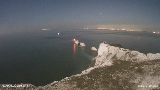 A night view of a shoreline with white cliffs meeting the sea. The water reflects faint lights and there are three prominent rock formations jutting out from the coast. In the background, a distant city skyline is visible, illuminated by lights. A small boat is seen on the water, and a red light is shining from one of the rock formations.