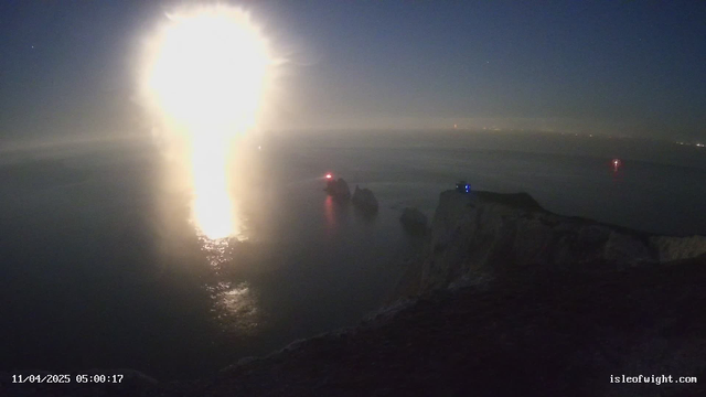 A coastal scene at night with a bright, glowing light reflecting off the ocean surface, creating a shimmering effect. In the distance, faint outlines of cliffs and rocks can be seen, with a few lights visible on the water, possibly from boats. The sky is dark with some stars visible. The date and time display at the bottom indicates it is April 11, 2025, at 5:00 AM.
