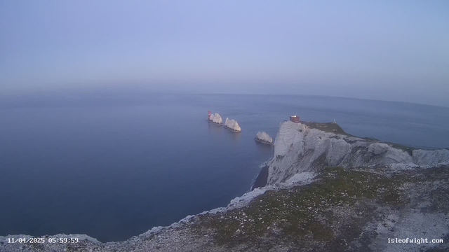 A coastal scene showing a rocky cliff overlooking calm waters. Three large, white rocks emerge from the water, with a red lighthouse structure on one of them. To the right, there is a small building on top of the cliff. The sky is light blue, and it appears to be early morning with soft, hazy light illuminating the scene. The ground near the cliff is covered in grass and small stones.