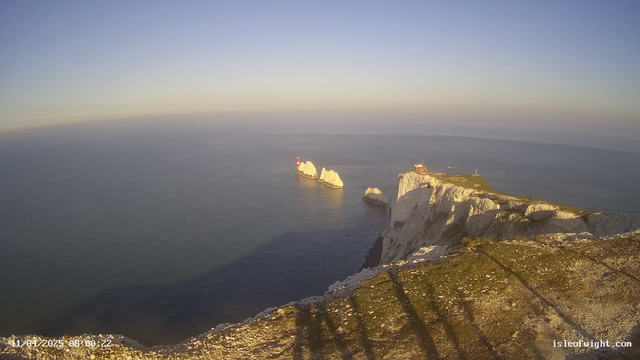 A scenic view of a coastal landscape featuring white cliffs and the ocean. The cliffs appear rugged, with some grassy areas at the top. In the distance, there are two prominent rocky outcrops in the water, one of which has a red lighthouse on it. The sky is clear with a soft gradient, transitioning from light blue to pale yellow near the horizon. The tranquil sea reflects the colors of the sky and is mostly calm. There are visible shadows cast by rocks in the foreground, suggesting the time is early morning.