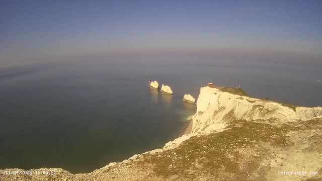 A panoramic view of a coastline featuring steep white cliffs meeting a calm blue sea under a clear sky. In the distance, there are three rocky outcrops with red and white lighthouses topped by a small building on the cliff edge. The foreground shows a grassy area with scattered stones. The date and time in the bottom left corner indicate 11 April 2023 at 11:00:51.