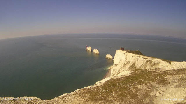 A scenic view of a coastline featuring white chalk cliffs that rise sharply from the sea. In the background, the calm blue water extends to the horizon under a clear blue sky. Some rocky formations and a small red and white lighthouse are visible in the water. The landscape includes a grassy area at the edge of the cliff, leading down to a sandy beach below.