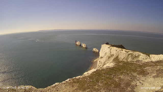 A panoramic view of the coast features white chalk cliffs rising sharply from the water. In the distance, a few rocky formations jut out into the sea. The water is calm and reflects the sunlight, creating a shimmering surface. The sky is mostly clear with a soft gradient of blue, indicating a sunny day. Along the cliff's edge, patches of grass and small rocks are visible, with a few people seen walking in the distance.
