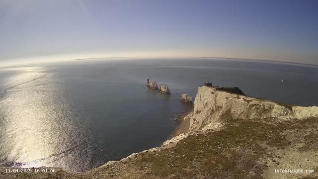 A panoramic view of a coastal landscape near a sea. In the foreground, there is a grassy cliff edge with a rocky surface. The cliff drops down towards the water, which is shimmering under sunlight. To the right, several large white rock formations emerge from the sea, with a hint of land visible in the distance. The sky above is clear and bright, with a gradient of light blue. In the upper left corner, a faint silhouette of distant land can be seen. The time and date are displayed at the bottom left corner of the image.