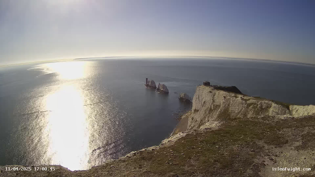A panoramic view of a coastline under a clear blue sky. The sun reflects brightly on the calm sea, creating a shimmering pathway across the water. In the foreground, there is a grassy cliff with some rocky edges. To the right, a small, dark structure is visible on top of the cliff. In the water, there are two white rock formations, with a ship nearby, adding a touch of activity to the serene landscape.