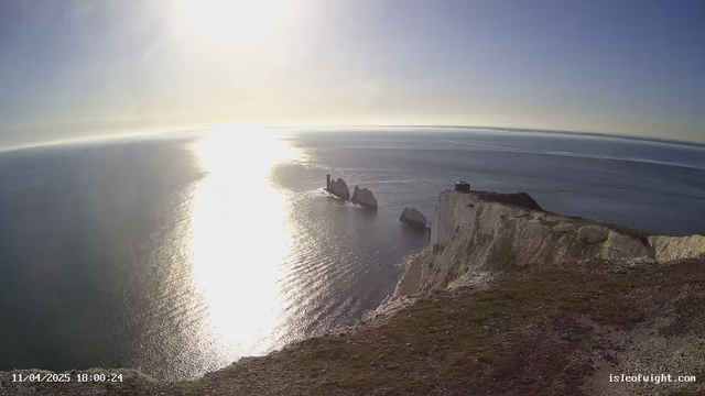 A panoramic view of the ocean at sunset, reflecting sunlight that creates a shimmering pathway on the water. In the distance, two rocky outcrops protrude from the sea, with a faint tower-like structure on one. The foreground features a grassy cliff edge leading into the water. The sky is clear, transitioning from blue to a lighter hue near the horizon where the sun sets. The bottom left corner displays the date and time stamp.