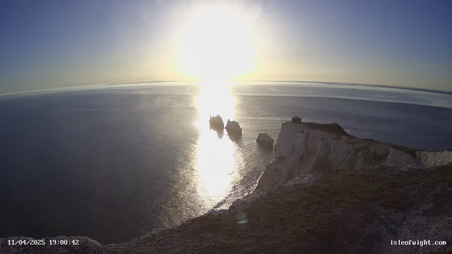 A scenic view of the ocean at sunset, with the sun shining brightly, casting reflections on the water's surface. There are three large rock formations jutting out of the water, and a cliff with a small building is visible on the right side. The sky transitions from deep blue at the top to lighter hues near the horizon, creating a tranquil atmosphere.