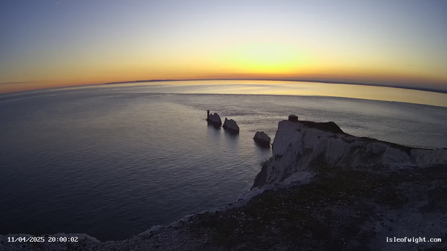 A scenic view of the ocean at sunset, with soft hues of orange and blue mixing in the sky. In the foreground, a steep, rocky white cliff reveals several prominent rock formations jutting out into the calm water. A structure, possibly a tower, is situated on one of the rocks. The water reflects the colors of the sky, creating a serene atmosphere. In the background, a distant shoreline can be seen.