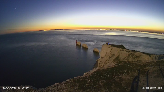 A scenic view featuring rocky cliffs at dusk. In the foreground, a rugged white cliff edge leads down to calm waters. Two recognizable rock formations protrude slightly from the water, one with a red lighthouse atop it. The sky transitions from deep blue to vibrant orange and yellow near the horizon, where distant lights from the coast are visible. The scene captures a tranquil ambiance as night approaches.