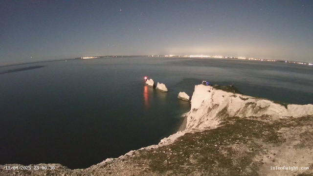 A serene nighttime view of a coastal landscape featuring large white chalk cliffs that descend into a calm sea. In the foreground, there are two prominent rock formations emerging from the water, with a faint red light visible on one of them. The sky is dark with scattered stars, and a distant shoreline illuminated by city lights can be seen in the background, indicating the presence of a nearby town or settlement. The overall atmosphere is tranquil and slightly mysterious, with the water reflecting the soft glow of the lights.