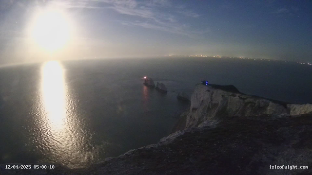 A serene coastal scene featuring a moonlit ocean. The bright moon reflects off the water, creating a shimmering path. In the lower right corner, a rocky cliff can be seen, partially illuminated. There are two distinct rock formations emerging from the water, with one displaying a red light on top. The horizon in the distance is softly lit, possibly indicating a nearby city. The sky is clear with a few wispy clouds, enhancing the tranquil atmosphere. The timestamp at the bottom indicates the image was captured on December 4, 2025, at 5:00 AM.