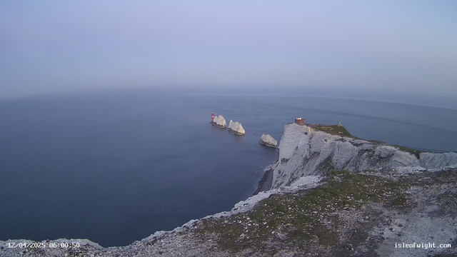 A coastal scene featuring white chalk cliffs and several rocky outcrops in the ocean. In the background, a red and white lighthouse stands on one of the rocks, while a small red building is visible on top of the cliffs. The sky is pale blue, and the sea appears calm and slightly reflective. The foreground includes some grassy areas on the cliff edge.