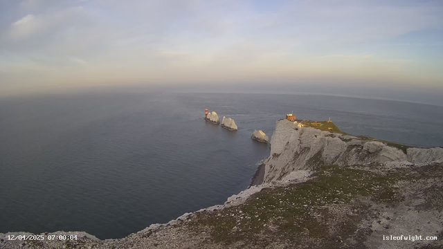 A coastal view featuring a rocky cliff edge on the right, with patches of green grass and gravel. Below the cliff, three prominent white chalk formations emerge from the ocean, with a red lighthouse visible on the leftmost rock. In the background, the sea transitions into a lighter blue towards the horizon, under a pale sky with soft clouds. A small, white building is situated on the cliff, along with some structures visible at the top of the cliffs towards the left.