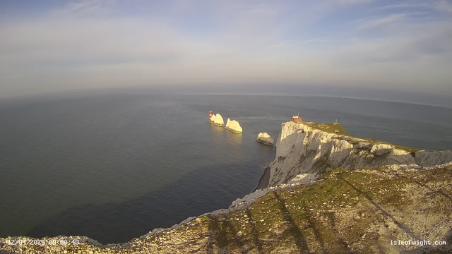 A scenic view of the coastline featuring white chalk cliffs meeting the blue sea. In the water, there are two prominent rock formations with a red lighthouse on one of them. The sky is partly cloudy with a soft blue hue, and the shoreline is grassy, leading to the edge of the cliffs.