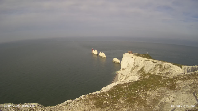 A coastal view featuring steep white chalk cliffs that descend into a calm sea. In the distance, there are two distinct rocky formations jutting out of the water, with a red lighthouse positioned on one of them. The sky above is mostly cloudy, and the scene conveys a peaceful, natural landscape with a subtle color palette of blues and greens. The foreground shows grassy terrain with rocky outcrops.
