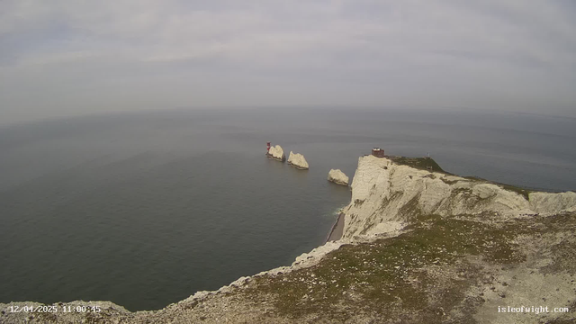 A coastal view features white chalk cliffs on the right side, descending towards the water. In the distance, two prominent rock formations extend into the sea, with a red and white lighthouse positioned on one of the rocks. The ocean appears calm and grey under a cloudy sky, with soft, muted colors dominating the scene. The foreground includes grassy terrain, with some rocky patches visible.