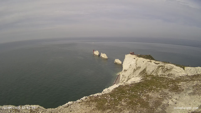 A coastal scene features white chalk cliffs descending into a calm sea. Three distinctive rock formations rise from the water, with a red and white lighthouse situated on one of the rocks. In the background, a small boat can be seen moving across the water. The sky is overcast with a light gray hue. The foreground has grassy patches with rocky outcrops, adding texture to the landscape.