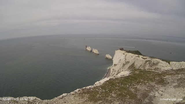 A seaside view featuring white cliffs on the right side, leading down to a rocky shore. In the water, there are several small boats and a red lighthouse located on a rock formation. The sky is overcast with gray clouds, and the ocean appears calm with gentle ripples. The scene captures a sense of tranquility and nature’s beauty along the coastline.