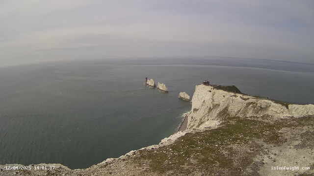 A coastal scene showing white cliffs along the edge of the sea. In the distance, several rock formations rise from the water, with one featuring a red structure on top. The sky is overcast with soft gray clouds, and the sea appears calm with gentle waves. The foreground includes green grass on the cliff, while the shoreline below has a small beach.