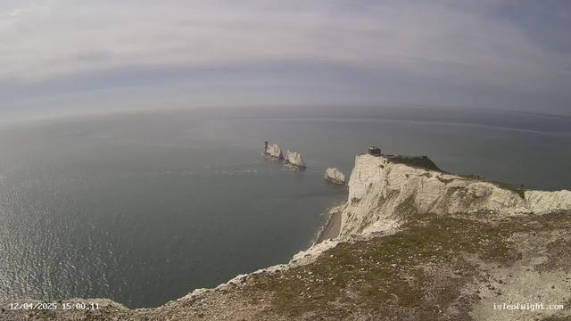 A scenic view of coastal cliffs meeting the ocean. The cliffs are white and jagged, with gentle slopes leading to the sea. In the distance, a series of rock formations is visible, with a lighthouse on one of them. The sky above is mostly clear with some clouds, and the water reflects light, creating a shimmering effect. The foreground shows grassy terrain with scattered stones.