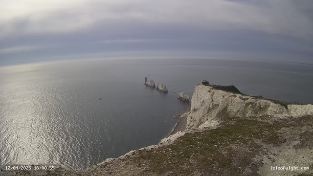 A coastal view features a high, rugged white chalk cliff on the right side. Below the cliff, there is a sandy beach leading to a calm, reflective sea. In the distance, two prominent rock formations rise from the water, while a lighthouse is visible atop one of the structures. The sky is partly cloudy, casting a soft light over the scene.